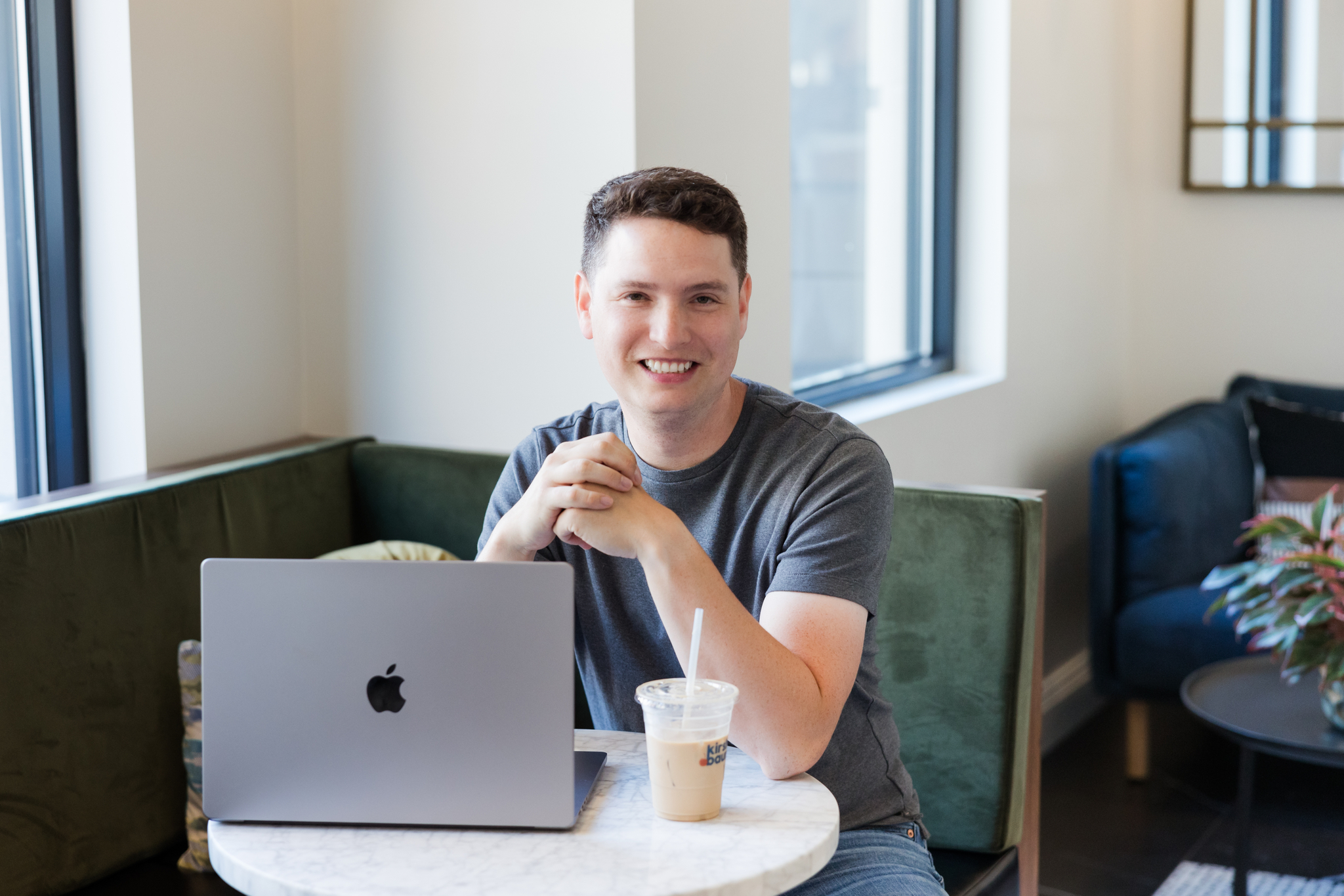 Justin Herrera at a laptop with an iced coffee, smiling, in a window-lit cafe.