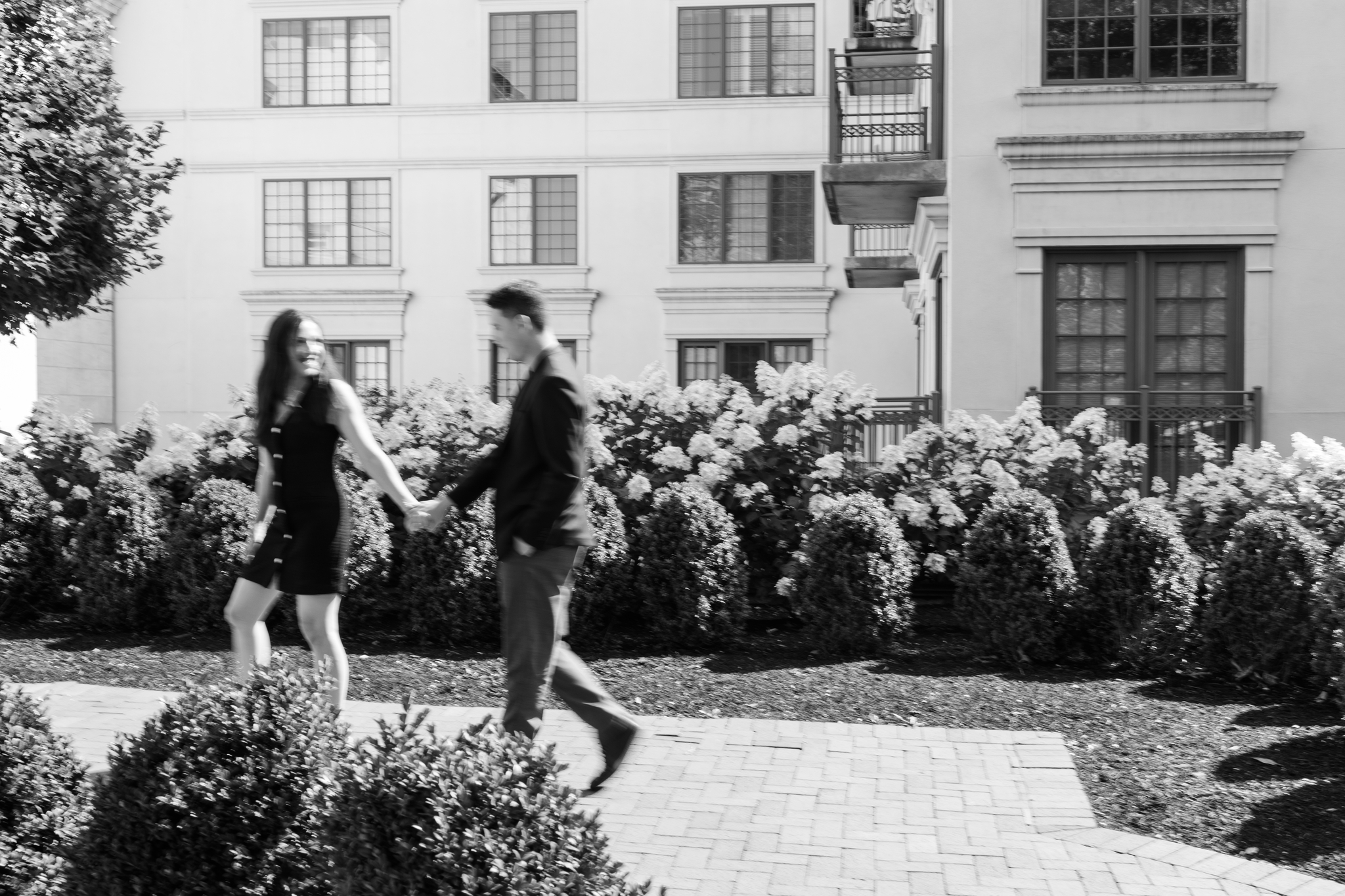 Black-and-white still from a wedding film — a couple walking hand-in-hand past hydrangeas.